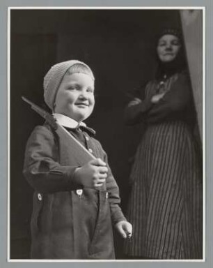 Portrait eines Jungen mit einem Holzgewehr und einer Frau im Hintergrund stehend