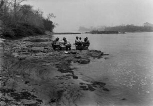 Ufer des Rio Grande : Männer am Ufer des Rio Grande. Zwei hocken neben einem Stapel, weitere in einem kleinen Boot. Im Hintergrund Flussinseln und Bäume am Ufer.