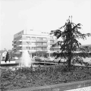 Rathausvorplatz: Blumenbeet mit Narzissen und Nadelbaum, Springbrunnen, Frau: Reeshoop [später Manfred-Samusch-Straße], Autos, Straßenlampen: hinten Wohn- und Geschäftsblock der nördlichen Randbebauung von Rathausplatz, mit Geschäften Schuhhaus "Bata", "Johs. Schmidt Markt"