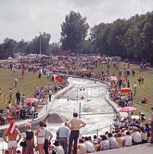 Hansjakob, Gottfried; Augsburg - Spickel, Am Eiskanal 30A; Kanuslalom Olympiastrecke, Freiflächengestaltung - Kanustrecke, Kanute beim Training mit Slalomtoren, Tribünen mit Zuschauern