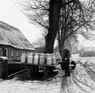 Person mit angelieferten Milchkannen (links), links ein Wohngebäude, Winterbild