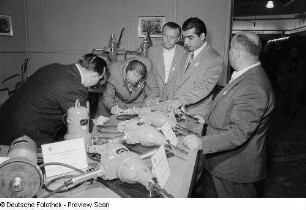 Eine schreibende Person umringt von weiteren Personen an einem Stand der Leipziger Herbstmesse 1954 mit ausgestellten Bohrmaschinen