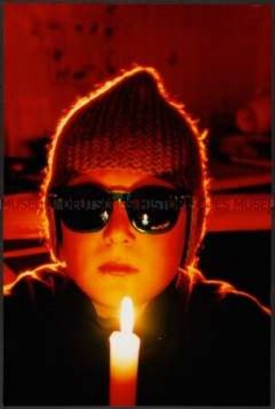 Kind mit Sonnenbrille und Mütze vor einer Kerze (Altersgruppe bis 14)