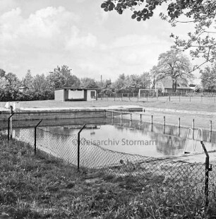 Freibad: Schwimmbecken: davor beschädigter Maschendrahtzaun: hinten links Verwaltungsgebäude: hinten rechts Sportplatz: im Hintergrund Bäume und Haus