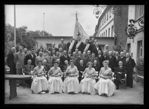 Gruppenbild Gesangverein Frohsinn Wertheim mit Standarte. Frauen in Abendkleid und Schärpe, Männer im Anzug und z.T. mit Schärpe. Im Hintergrund Hotel "Zum Schwanen", Männer sitzend: 1.v.r. sitzend Lokomotivführer Georg Zwirner, 3.v.r. sitzend Maurer und Vorstand Jakob Bodenschatz, 4.v.l. sitzend Sattler Otto Hodapp, 3.v.l. sitzend Spediteur Ernst Hoh, 2.v.l. sitzend Maler Karl Bodenschatz; Männer 1. Reihe stehend: 1.v.l. sitzend Kaufmann Ludwig Dreikorn; Männer 1. Reihe stehend: 1.v.l. stehend Buchdrucker Lorenz Dreikorn, 4.v.l. stehend Spengler Heinrich Schreck, 6.v.l. stehend Schreiner Adam Schmidt, 2.v.r. stehend Installateur Andreas Scherz, 1.v.r. stehend Maler Karl Oetzel, 2. Reihe stehend: 2.v.r. stehend Schmied Christoph Dinkel, 5.v.l. stehend evtl. Gemüsehändler Karl Seelig, letzte Reihe 1.v.l. stehend Glaser Karl Götz, 4.v.l. stehend Kontrolleur Karl Garrecht und 2.v.r. stehend Sandschöpfer Jakob Herz