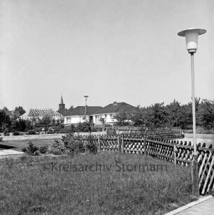 Willinghusener Straße: vorn Rasenflächen mit Jägerzaun: rechts Straßenlampe: hinten Einfamilienhäuser: links eines im Rohbau: im Hintergrund evangelische Kirche St. Johannis