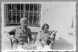 Hans und Etha Richter, im Freien vor einem vergitterten Fenster an einem Tisch sitzend