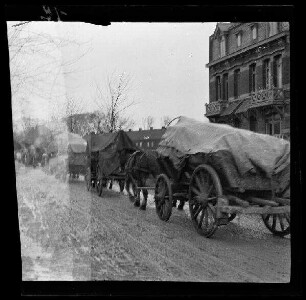 Nicht identifizierbare Ortschaft in Nordfrankreich oder Belgien: Pferdegespanne mit Planwagen auf einer morastigen Ortsstraße (Rückansicht)