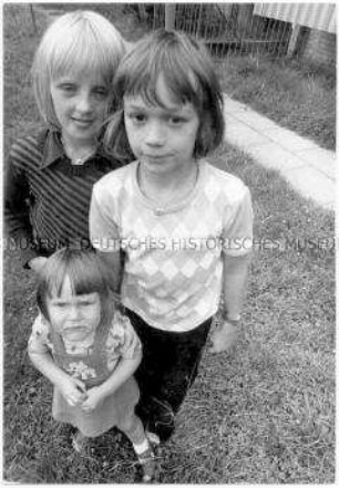 Drei Mädchen, das kleinste steht vor den beiden größeren, eines streckt die Zunge heraus