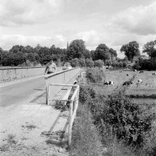 Trave: Brücke der Landstraße Richtung Reecke (Hansestadt Lübeck): links Mann: dahinter Pkws: hinten rechts Weide mit Rinderherde: im Hintergrund Kirchturm zwischen Bäumen