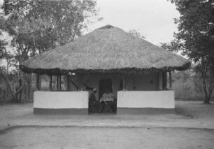 Der Sultan : Blick auf ein Haus in Landschaft. Expeditionsmitglieder sitzen mit einem Dorfbewohner am Tisch.