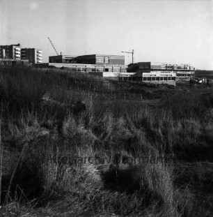 Schule am Masurenweg [Dörfergemeinschaftsschule, Grund- und Hauptschule]: Rohbau: links Haus und Wohnblocks: dahinter Baukräne: vorn Wiese, Zäune, Erdhaufen: 11.11.1972
