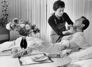 Werbefotografie für einen Prospekt der Techniker Krankenkasse (TK). Das Model, Frau Wagner, betreut einen an Grippe erkrankten Mann. Fürsorglich legt sie ihm einen Schal um den Hals