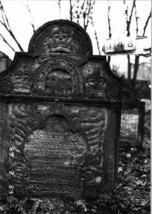 Grabstein 148 (Index: 99751968) Material: Sandstein Zustand: stark verwittert mit Textverlust Maße HxBxT: 142x99x10 cm Beschreibung: Stele, in Voluten auslaufend. Rundbogenaufsatz, Schriftfeld in Wappenschildform Symbolik: drei Kronen: Grabstein 148