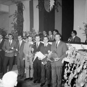 Kreisfußballverband Stormarn: Verbandstag: Reinfeld: Festsaal des "Hotel Stadt Hamburg": Ehrung der Meister: Geehrte mit Blumen, Ehrenteller: dahinter Verbandsleitung auf Podium: oben Wappen Schleswig-Holstein, Grün- und Blumenschmuck, 22. Mai 1965