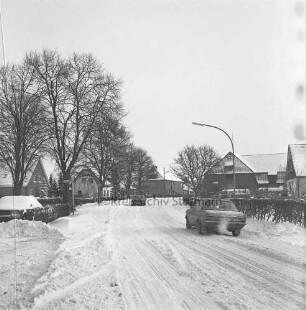 Hauptstraße, Landesstraße L89: Pkws: Häuser, Jägerzaun, parkender Pkw, Gartenhecken, Straßenbäume, Straßenlampe: links Abzweig Alte Dorfstraße: vorne links zusammengeschobener Schnee: im Winter