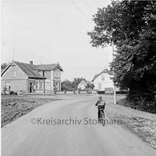 Rohlfshagen: Ortseingang: vorn Straße aus Richtung Barkhorst: rechts Schulkind auf Fahrrad: rechts dahinter Ortseingangsschild: daneben Bäume: hinten links Häuser