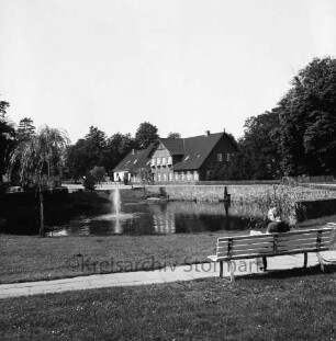 Dorfstraße: Dorfteich mit Springbrunnen: vorn Grünanlage: rechts Bank mit Frau: hinten Häuser und Bäume