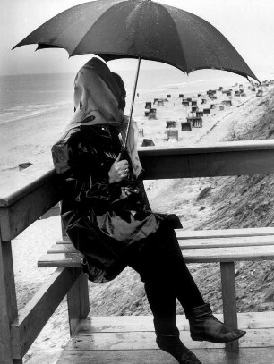Insel Sylt. Ferien. Eine Frau sitzt an einem verregneten Tag mit einem Regenschirm auf einer Bank an einem Aussichtspunkt. Kein Mensch befindet sich am Strand
