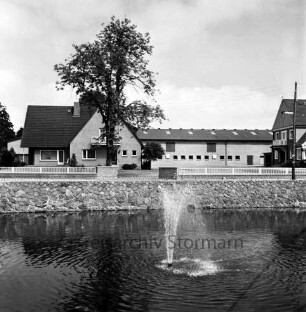 Dorfstraße: vorn Dorfteich mit Springbrunnen: hinten Häuser und Baum