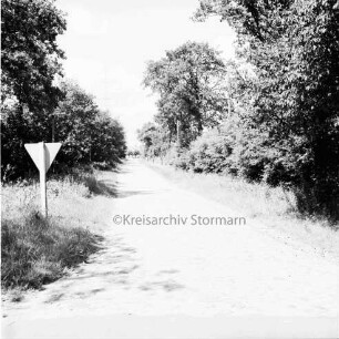 Feldweg: links Verkehrsschild: rechts Strommasten: links und rechts Büsche und Bäume