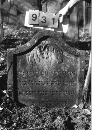 Grabstein 931 (Index: 99753183) Material: Sandstein Zustand: leicht verwittert mit Textverlust Maße HxBxT: 71x64x7 cm Beschreibung: Stele, Konvexbogenabschluß, Schriftfeld eingetieft Symbolik: nach unten hängende Palmzweige: Grabstein 931