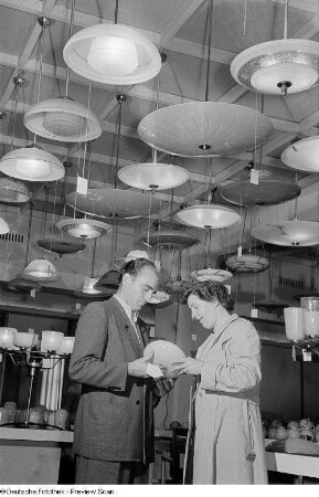 Ein Mann und eine Frau begutachten einen gläsernen Lampenschirm, im Hintergrund eine Vielzahl ausgestellter Deckenlampen der Leipziger Herbstmesse 1954