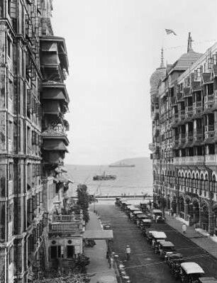 Bombay (Indien). Straßenflucht mit parkenden Automobilen und Taj-Mahal-Hotel. Blick zum Hochseepassagierdampfer "Cleveland"