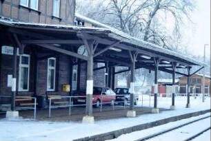 überdachter Bahnsteig mit parkenden Autos; hinten ehemalige Toilettenhäuschen