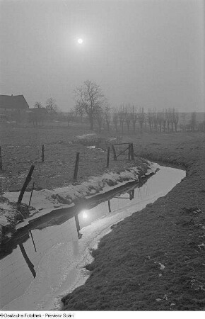 Ein vereister Bachlauf mit sich spiegelnder Sonne, im Hintergrund kahle Bäume und Häuser