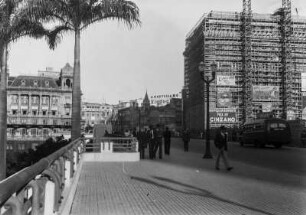 Straßenszene : Blick auf eine belebte Straße mit Passanten. Rechts ein eingerüstetes Gebäude mit Werbeschildern, im Hintergrund weitere Gebäude.