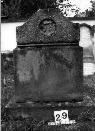 Grabstein 29 (Index: 99804572) Material: Sockel Sandstein, Aufsatz Kalkstein ? Zustand: Stele mit Inschrift fehlt Maße HxBxT: 96x60x24 cm Beschreibung: getreppter Sockel, giebelartiger Aufsatz mit flachem Abschluß: Grabstein 29