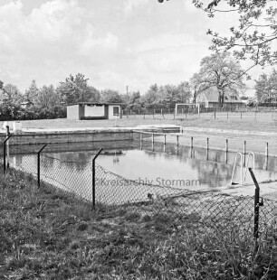 Freibad: Schwimmbecken: davor beschädigter Maschendrahtzaun: hinten links Verwaltungsgebäude: hinten rechts Sportplatz: im Hintergrund Bäume und Haus
