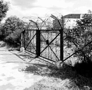 Glinde: Oher Weg: Gelände des ehemaligen Heereszeugamtes: vorn Metalltor mit Stacheldraht: rechts und links Büsche: hinten mehrere Hallen