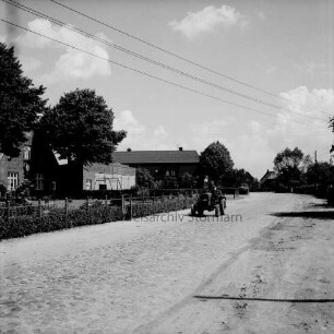 Hauptstraße mit Kopfsteinpflaster: Trecker: dahinter Bauernhäuser mit Vorgärten und Hecken
