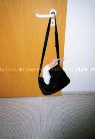 An einer Türklinke hängende Handtasche mit einem künstlichen Arm (Altersgruppe bis 14)