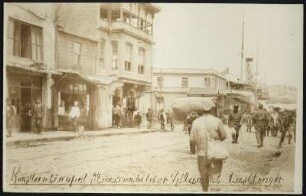 Belebter Straßenzug mit alten Geschäftshäusern in Istanbul nahe des Bosporus-Ufers, im Vordergrund Lastenträger, im Hintergrund Schiffe und Blick auf das gegenüberliegende Ufer