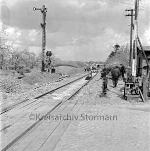 Bahnübergang bei Schrankenposten 75, an B 75: Bahnstrecke zwischen Bad Oldesloe und Neumünster: Gleiserneuerung: Ausrichten von Abstand der neuen Schienen mit Spezialfahrzeug: Arbeiter liegt im Gleisbett, Arbeiter und Bahnbeamte als Beobachter: links Schienenstücke, Signalanlage: vorne Bahnübergang, rechts Schranke geöffnet, Handkurbel, Schrankenwärterhäuschen, Strommast