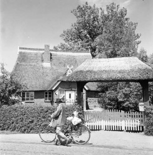 Hauptstraße: das alte Postgebäude mit Hecke und Tor, davor ein Fahrradfahrer