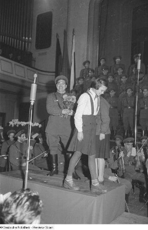 Eine Pionierin übergibt einem bulgarischen Soldaten einen Blumenstrauß, im Hintergrund Militärchor und -orchester eines bulgarischen Ensembles