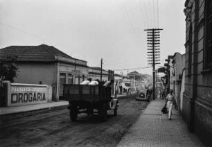 Straße in Olimpia : Straßenszene in Olimpia. Rechts und links der Straße Gebäude. Auf der Straße Fahrzeuge, rechts ein Passant auf dem Bürgersteig und Strommasten am Straßenrand.