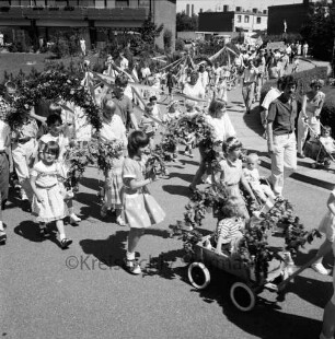 Kindervogelschießen und Dorffest: Festumzug: Kinder mit Blumenkranzbögen und geschmückten Bollerwagen: rechts am Straßenrand Zuschauer: im Hintergrund Reihenhäuser und Schornsteine der Paech-Brot GmbH