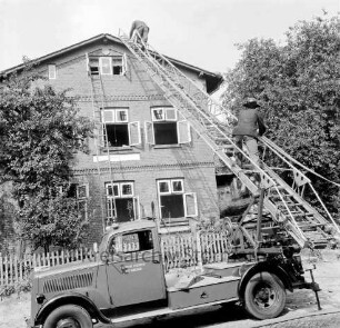 Freiwillige Feuerwehr Bad Oldesloe: Brandeinsatz: Wohnhaus mit geöffneten Fenstern, angelehnte Holzleiter: Leiterwagen mit ausgefahrener Drehleiter, zwei Feuerwehrmänner klettern auf Hausdach: Lattenzaun, Bäume