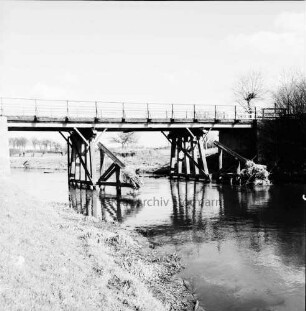Trave: Holzbrücke in baufälligem Zustand: davor Pfeiler mit Treibgut: rechts Büsche: hinten Wiesen und Bäume