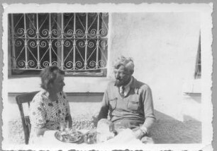 Hans und Etha Richter, im Freien vor einem vergitterten Fenster an einem Tisch sitzend