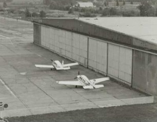 Flugzeughalle : Verkehrslandeplatz (ehemaliger Militärflugplatz). Blick vom Tower auf zwei Sportflugzeuge vor der Flugzeughalle (um 1960)