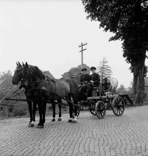 Freiwillige Feuerwehr Stubbendorf: Handdruckspritze von 1907 mit Pferdegespann: darauf zwei Feuerwehrmänner in Uniform: vorn Kopfsteinpflaster: hinten rechts Baum: hinten Häuser und Bäume