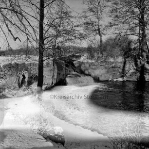 Mühlenteich mit Wehr und Überlauf: vorn Böschung: links und rechts Bäume: Winter