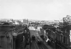 Straßenszene : Blick von einem Dach auf eine Straße, auf der eine Tram und ein Bus fahren. Im Hintergrund blick über die Stadt.