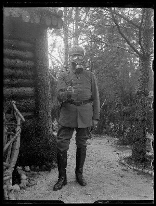 Unbekannte Ortschaft [Litauen/Weißrussland/Ukraine?]: Offizier mir Pickelhaube, aufgesetzter Gasmaske und vorgehaltener Pistole neben einem Holzhaus